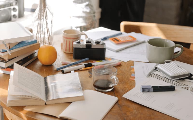 La photo montre une table de travail encombrée de nombreux cahiers, livres, stylos et d'un verre de café.