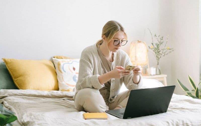 La photo montre une femme assise sur son lit, travaillant sur son ordinateur portable tout en utilisant son téléphone pendant le télétravail.