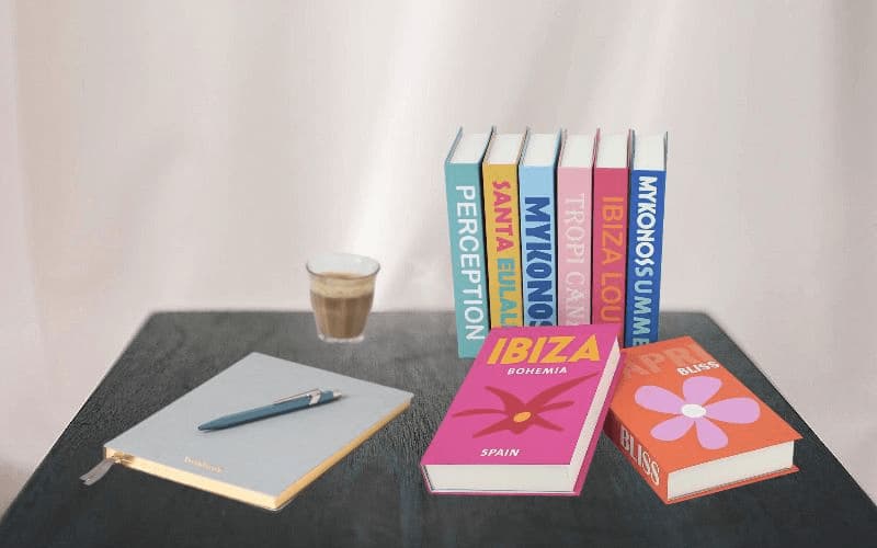 La photo montre plusieurs faux livres placés les uns à côté des autres sur une table. Un cahier et une tasse de café sont posés à côté.
